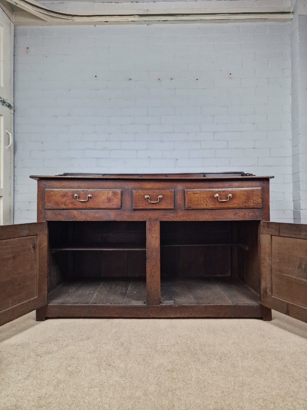 An antique dark oak dresser base featuring three upper drawers above two fielded panel cupboard doors with brass drop handles and a stepped upper gallery. View VII