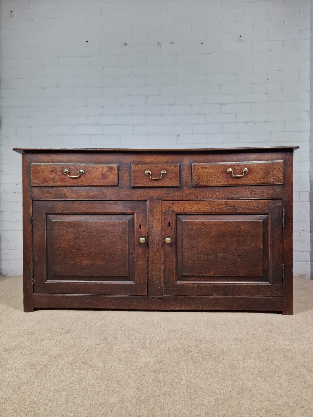An antique dark oak dresser base featuring three upper drawers above two fielded panel cupboard doors with brass drop handles and a stepped upper gallery. View VIII