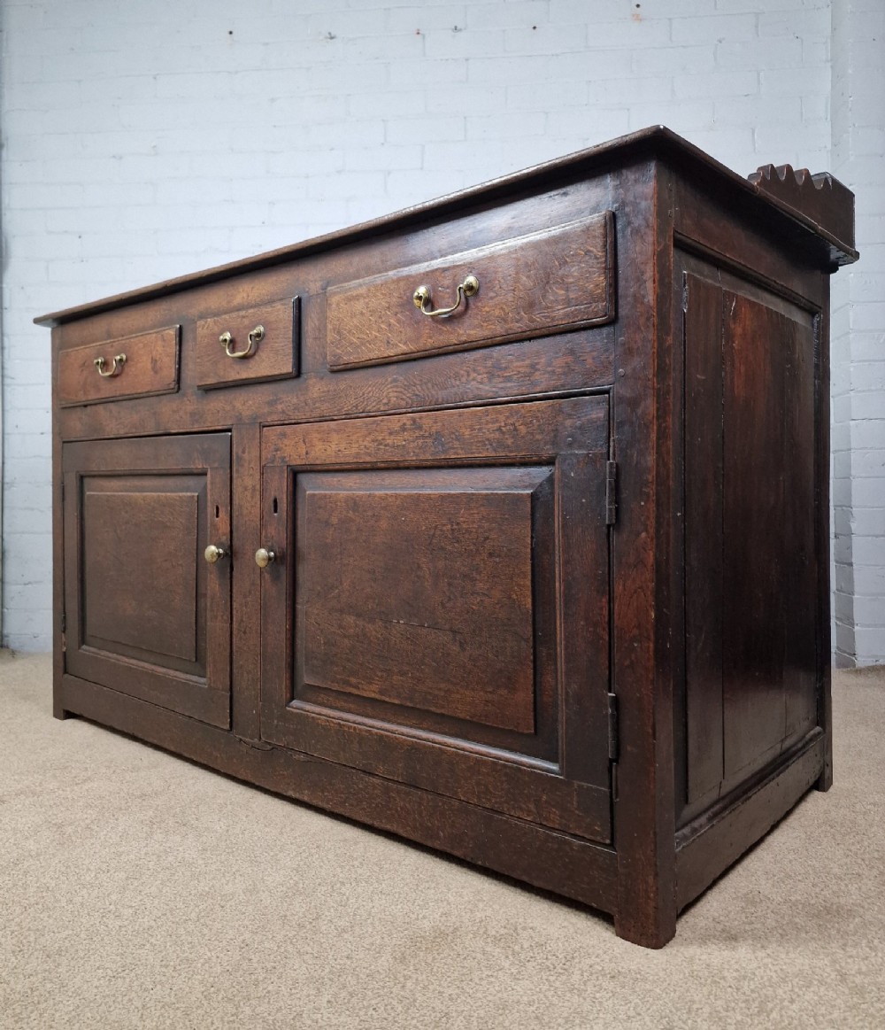 An antique dark oak dresser base featuring three upper drawers above two fielded panel cupboard doors with brass drop handles and a stepped upper gallery. View IX