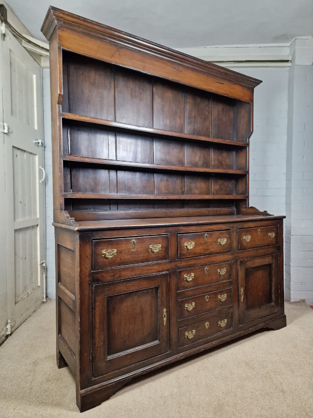 An antique dark oak dresser featuring a three-shelf open plate rack mounted above a base with multiple drawers and lower panelled cupboard doors. View IV