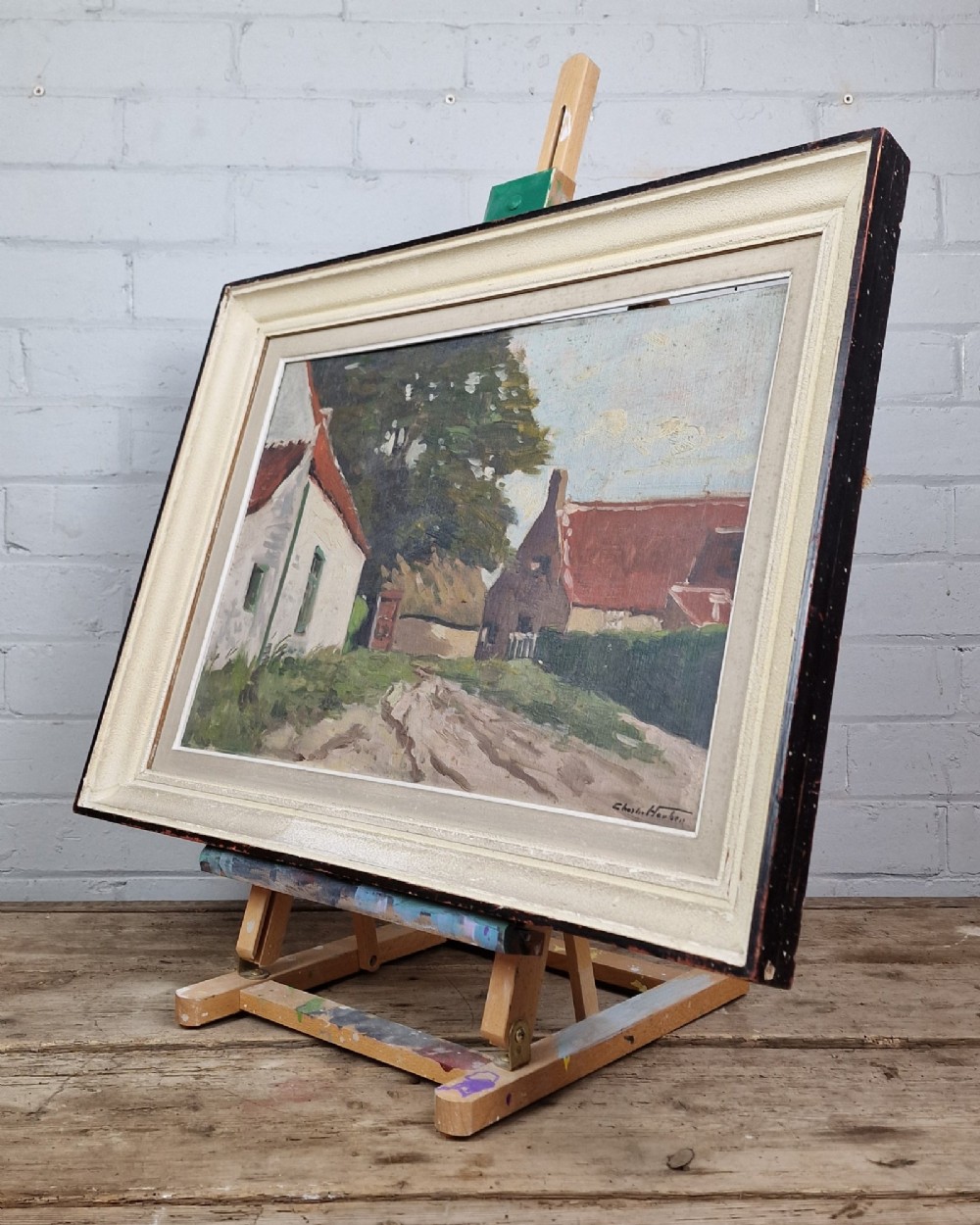 An oil painting on board depicting a Belgian farmyard scene with whitewashed cottages and trees, housed in a wide off-white molded frame. View V