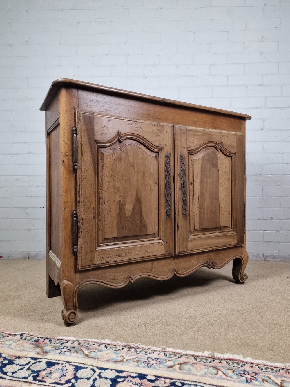 An antique French provincial two-door wooden side cabinet featuring carved panel doors, iron escutcheons, and short cabriole legs. View X