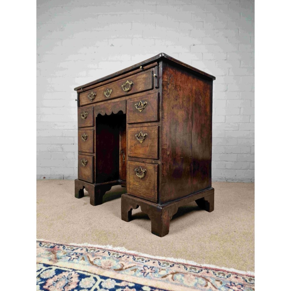 An early Georgian warm brown walnut kneehole desk featuring brass drop handles, side drawers, a central recessed cupboard, and bracket feet.