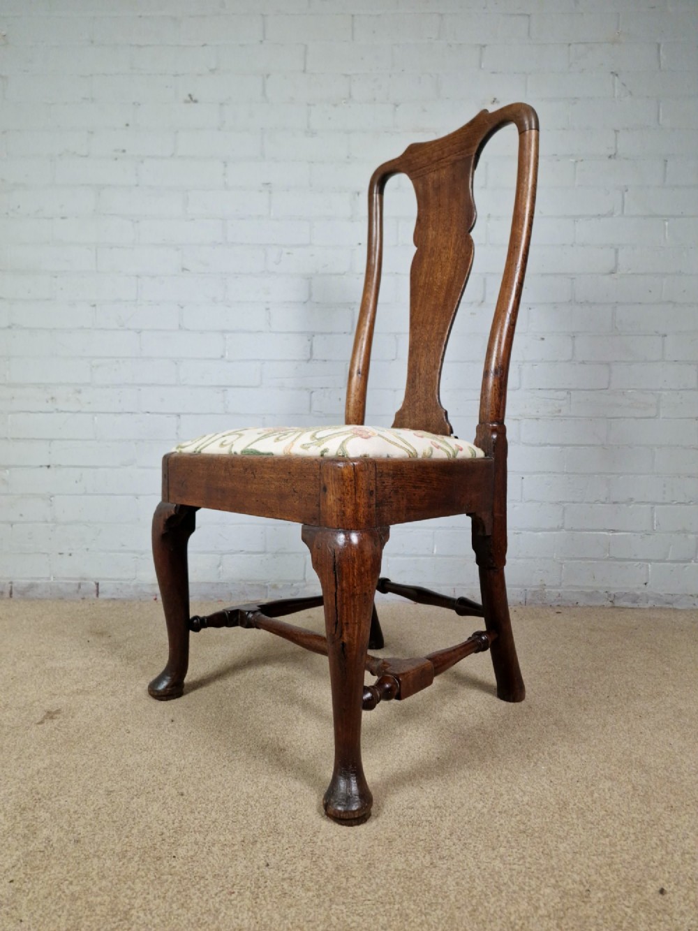 An early 18th century Queen Anne wooden dining chair featuring cabriole front legs, a vasiform back splat, and a patterned drop-in seat cushion.