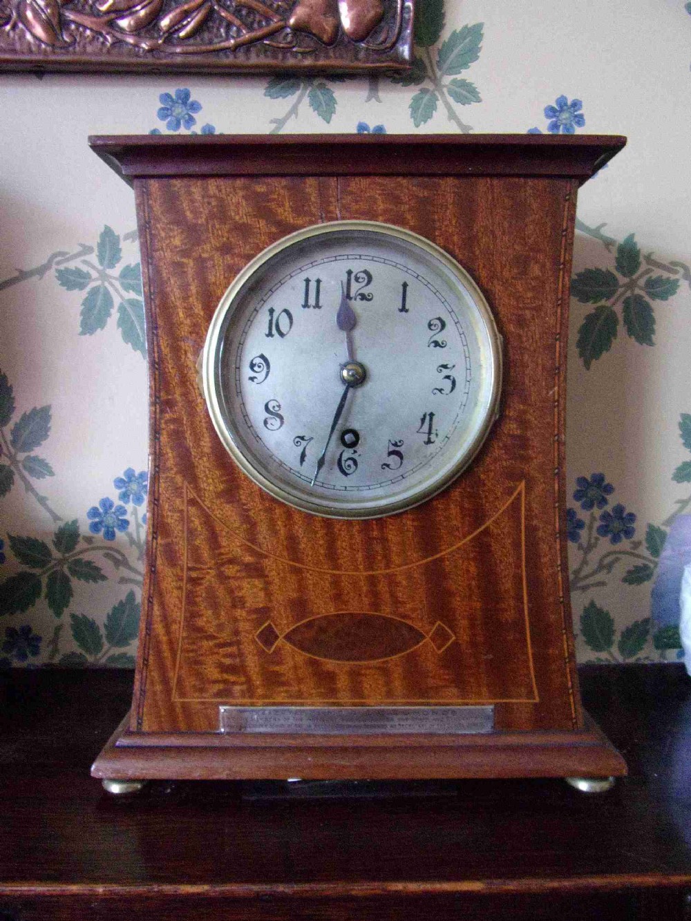 An Arts and Crafts Edwardian mahogany mantel clock featuring geometric line inlay, a circular white dial with Arabic numerals, and brass bun feet.