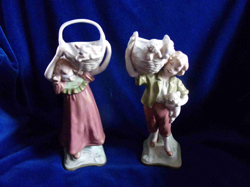A pair of early 20th-century Royal Dux porcelain figurines depicting a boy and a girl carrying baskets containing small animals.