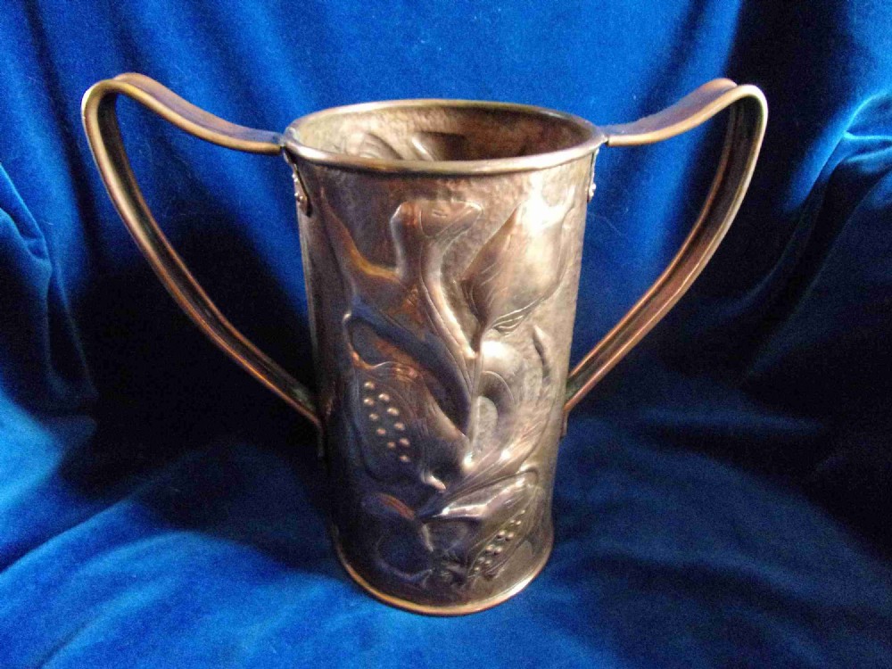 A hand-hammered cylindrical copper vase featuring two large loop handles and embossed botanical floral patterns in repousse relief.