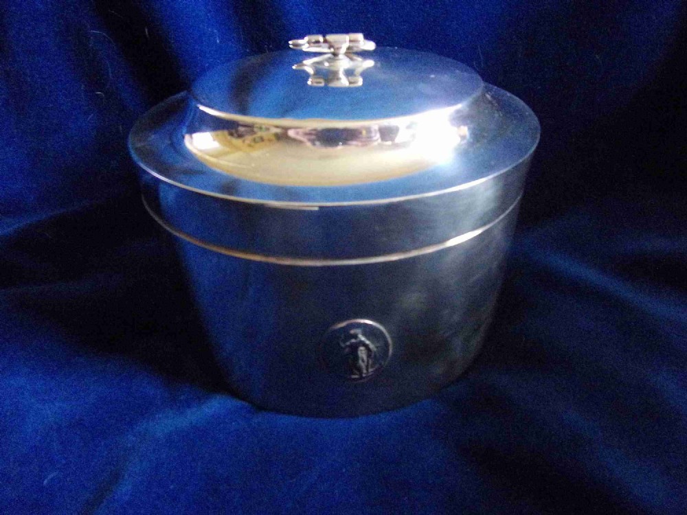 A cylindrical silver-plated lidded box featuring a circular relief emblem on the front and a loop finial handle on top.