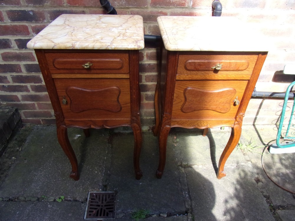A pair of French oak bedside cabinets featuring mottled beige marble tops, single drawers, cupboard doors with carved raised panels, and slender cabriole legs.