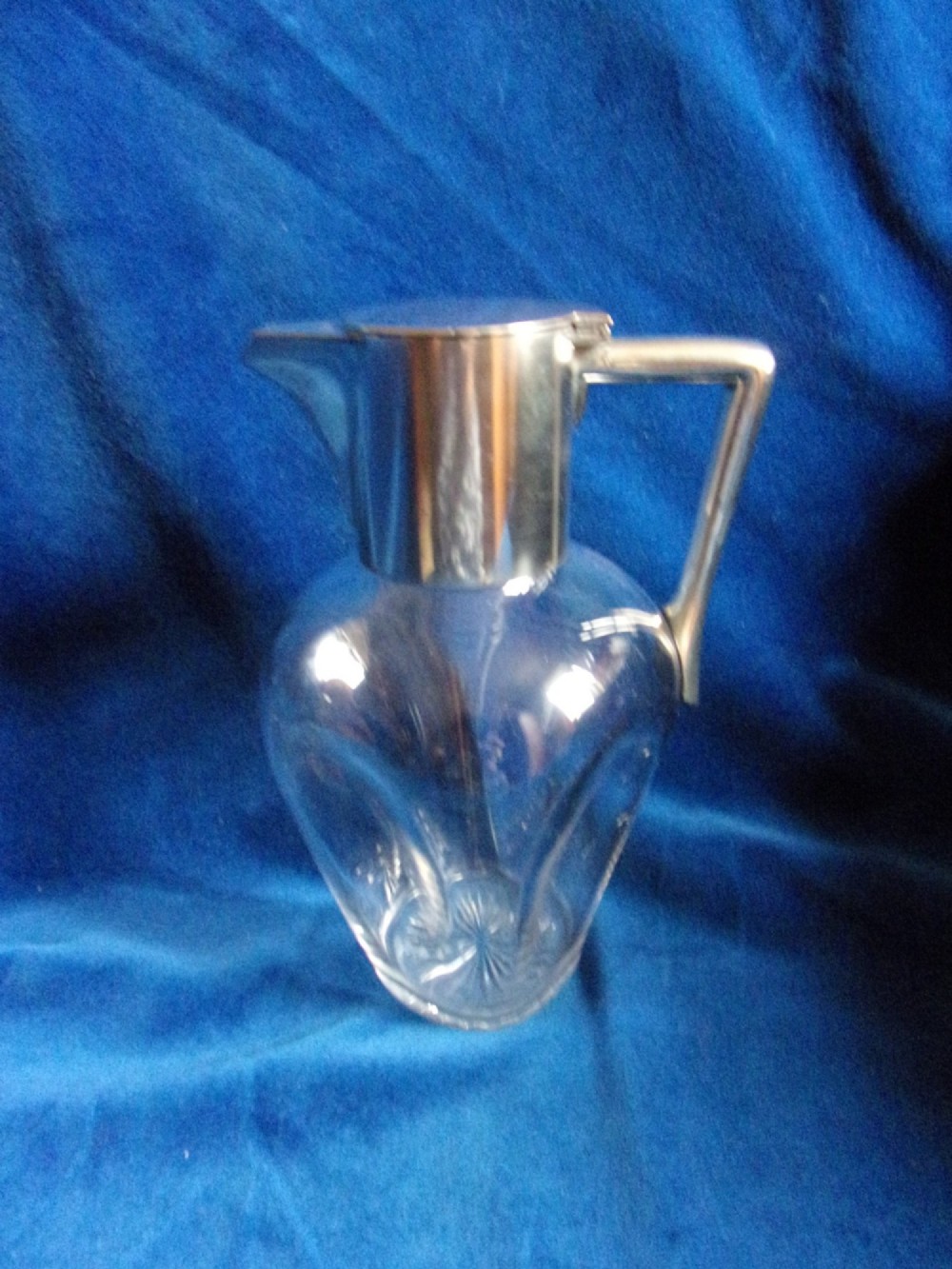 An Arts and Crafts clear glass claret jug featuring a silver-plated hinged collar, angular handle, and flat lid.