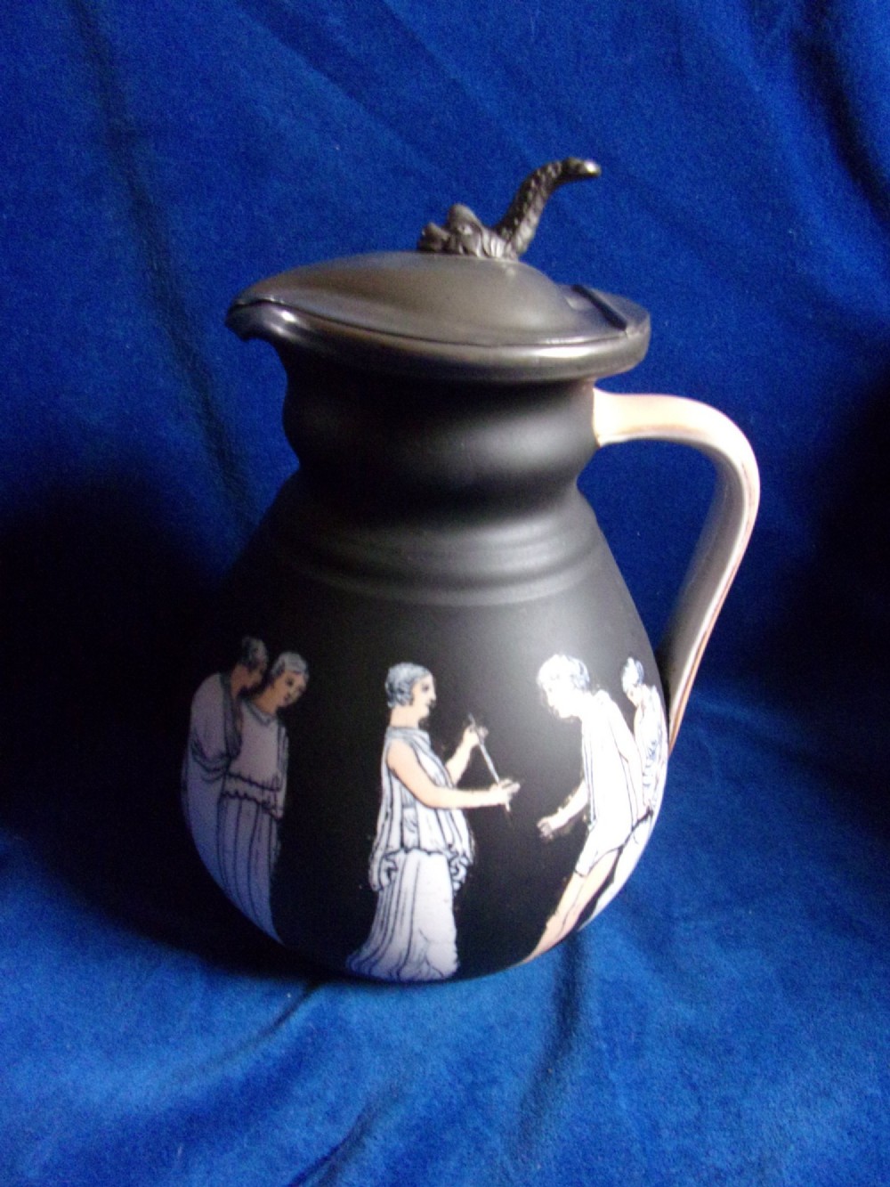 A matte black ceramic wine jug featuring classical white Greek figures around the body and a dark grey pewter hinged lid topped with a sculptured finial.