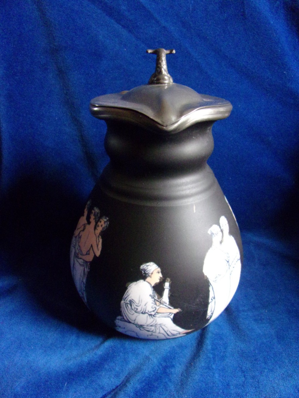 A matte black ceramic wine jug featuring classical white Greek figures around the body and a dark grey pewter hinged lid topped with a sculptured finial. View II