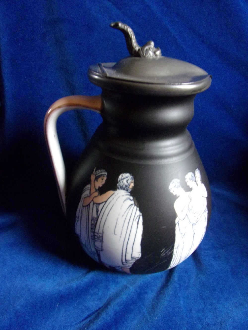 A matte black ceramic wine jug featuring classical white Greek figures around the body and a dark grey pewter hinged lid topped with a sculptured finial. View III