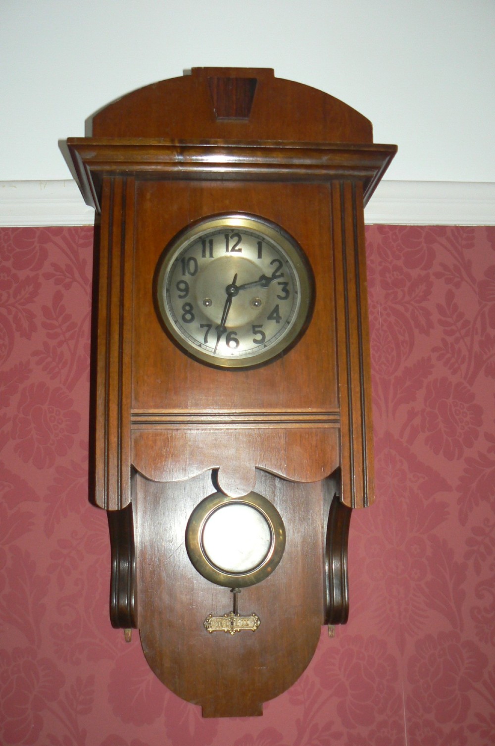An Arts and Crafts oak wall clock featuring a circular brass dial with Arabic numerals and an exposed circular brass pendulum aperture.
