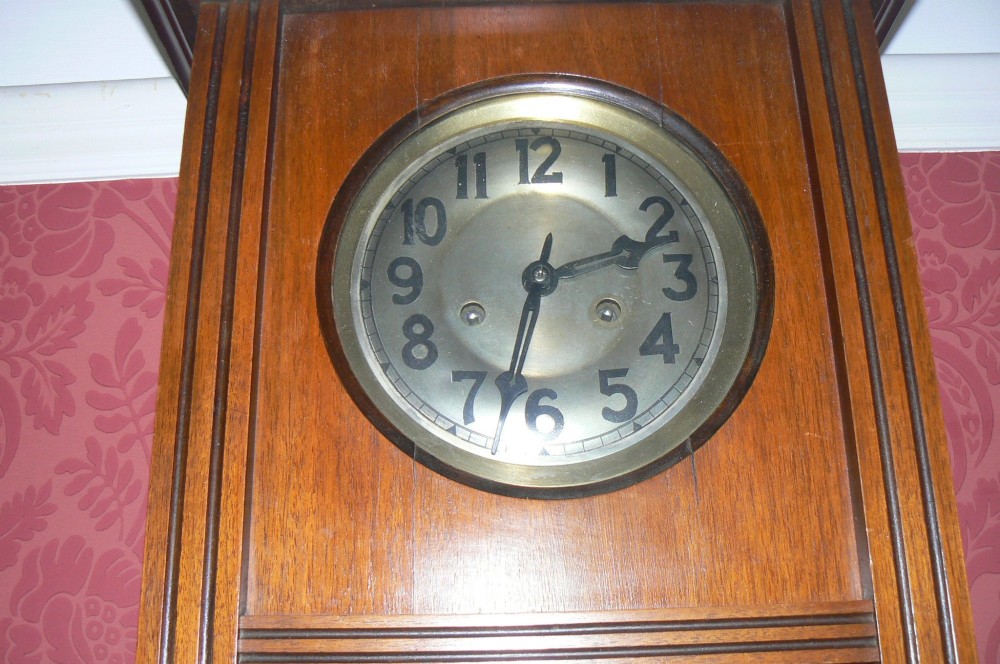 An Arts and Crafts oak wall clock featuring a circular brass dial with Arabic numerals and an exposed circular brass pendulum aperture. View II