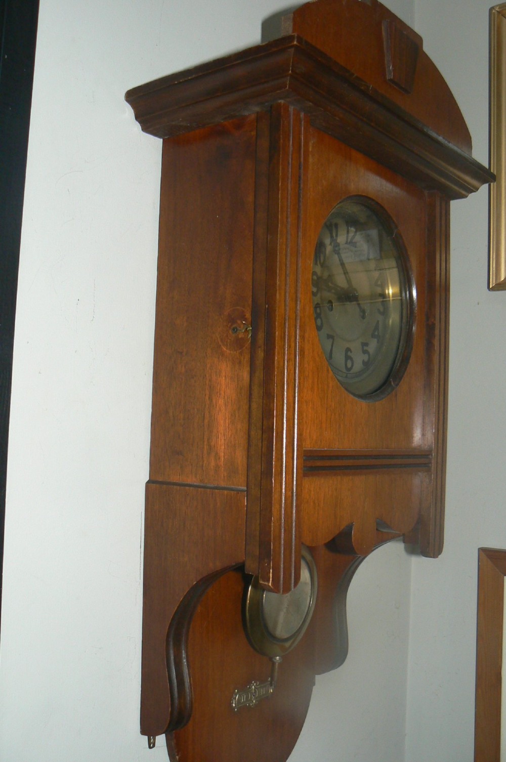 An Arts and Crafts oak wall clock featuring a circular brass dial with Arabic numerals and an exposed circular brass pendulum aperture. View IV