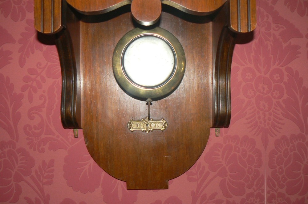 An Arts and Crafts oak wall clock featuring a circular brass dial with Arabic numerals and an exposed circular brass pendulum aperture. View V