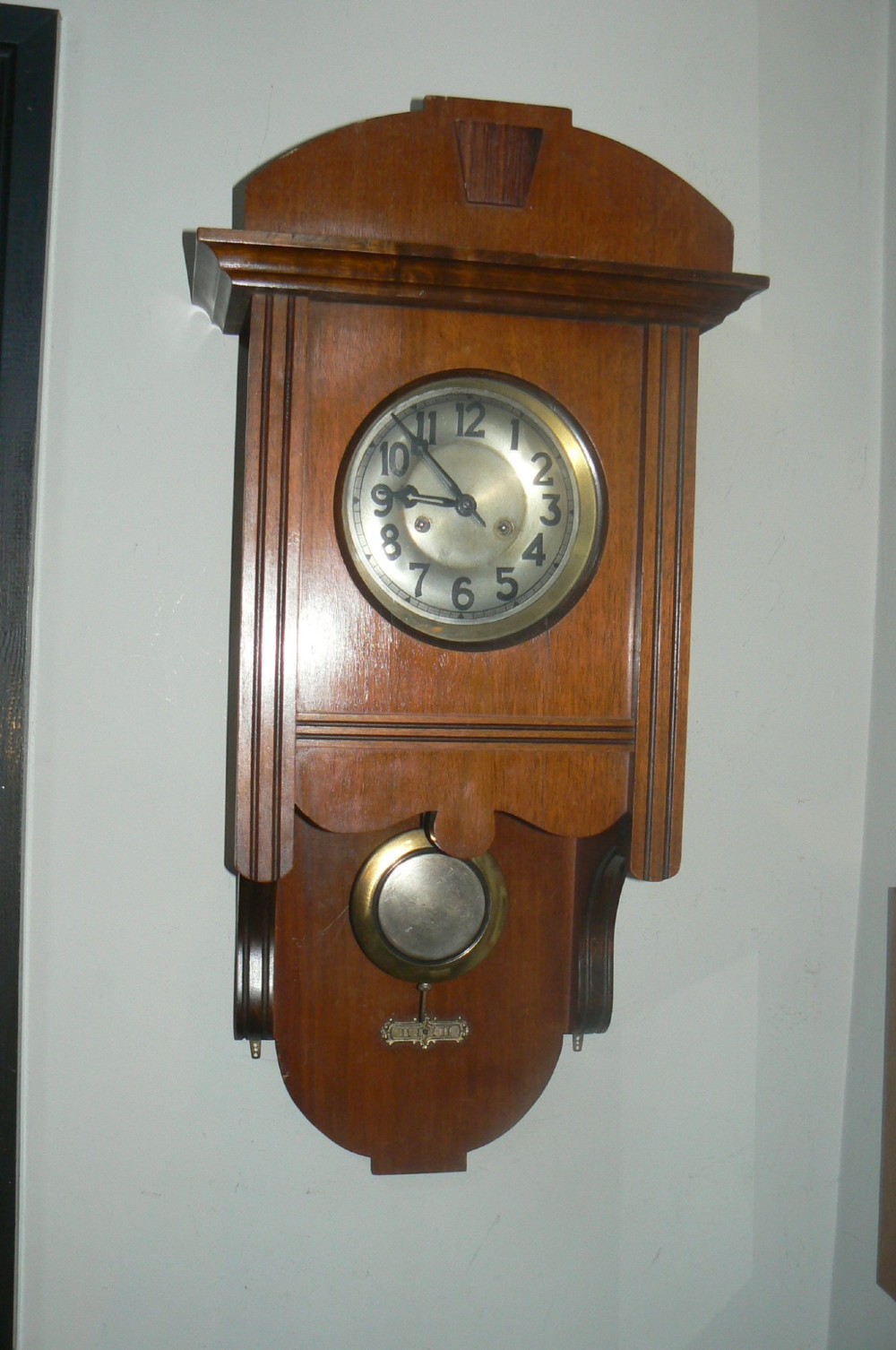 An Arts and Crafts oak wall clock featuring a circular brass dial with Arabic numerals and an exposed circular brass pendulum aperture. View VI