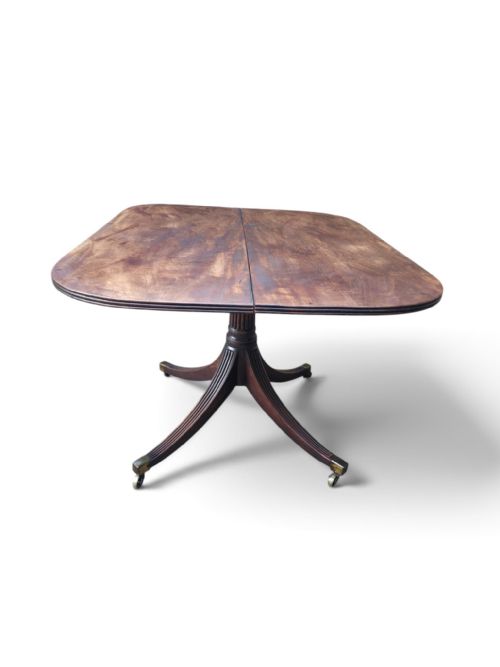 fig 2. An antique mahogany Regency style tilt-top dining table shown in its tilted vertical position, revealing its central reeded pedestal, splayed legs with brass castors, and underside brass latch mechanism.