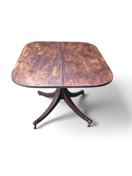 fig 3. An antique mahogany Regency style tilt-top dining table shown in its tilted vertical position, revealing its central reeded pedestal, splayed legs with brass castors, and underside brass latch mechanism.