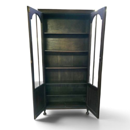 fig 1. An antique dark oak Arts and Crafts glazed bookcase featuring two tall glazed doors with arched panels above carved lower sections, supported on turned bun feet.