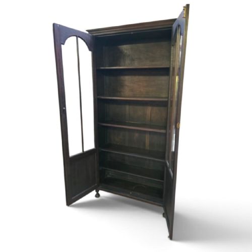 fig 2. An antique dark oak Arts and Crafts glazed bookcase featuring two tall glazed doors with arched panels above carved lower sections, supported on turned bun feet.