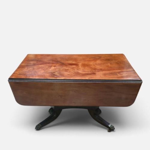 fig 1. A Regency mahogany drop leaf dining table featuring a flame mahogany top, two hinged side flaps, a central pedestal base, and reeded sabre legs ending in brass claw feet with castors.
