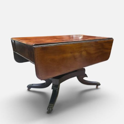 fig 2. A Regency mahogany drop leaf dining table featuring a flame mahogany top, two hinged side flaps, a central pedestal base, and reeded sabre legs ending in brass claw feet with castors.