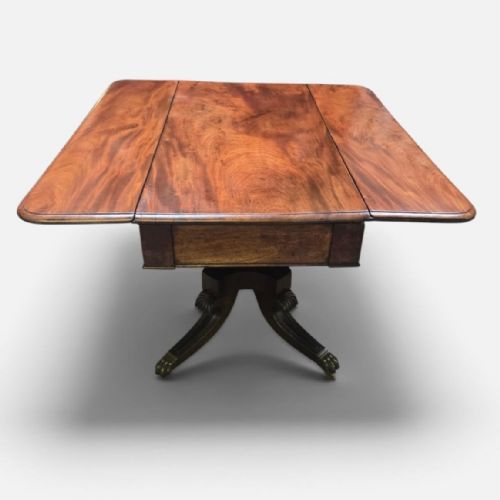 fig 3. A Regency mahogany drop leaf dining table featuring a flame mahogany top, two hinged side flaps, a central pedestal base, and reeded sabre legs ending in brass claw feet with castors.