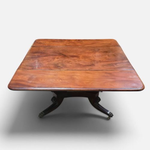 fig 4. A Regency mahogany drop leaf dining table featuring a flame mahogany top, two hinged side flaps, a central pedestal base, and reeded sabre legs ending in brass claw feet with castors.