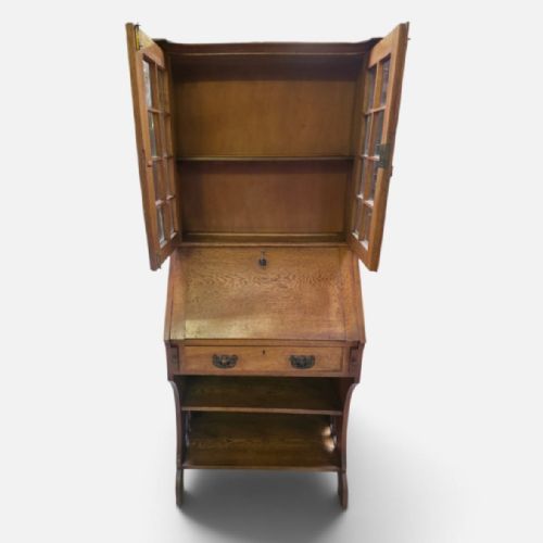 fig 1. An Arts and Crafts oak bureau bookcase featuring a twin-door glazed upper cabinet, a slant-front writing slope, a single drawer, and open shelving below.