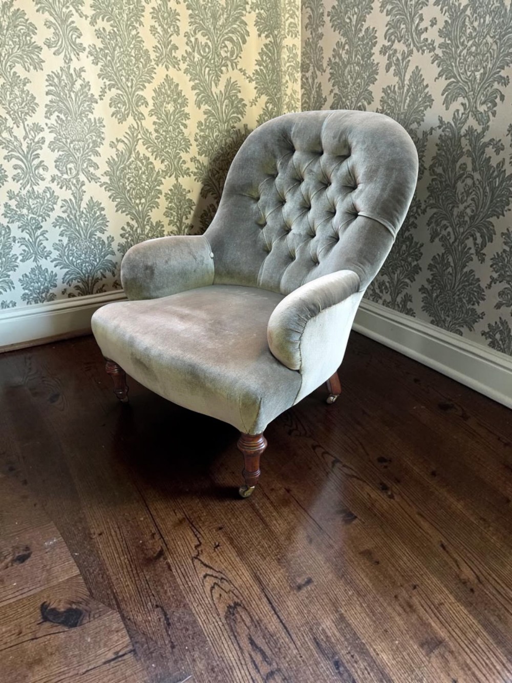 An Edwardian button-back armchair upholstered in light sage green velvet with turned wooden front legs on brass castors.