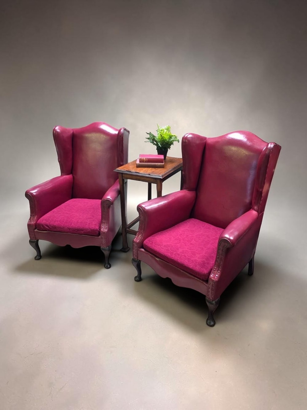 A pair of 1930s burgundy leather wingback armchairs featuring patterned fabric seat cushions, studded trim, and dark timber cabriole legs.