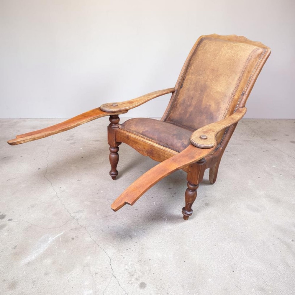 An antique 19th-century colonial planters chair featuring a reclined tan leather seat and backrest alongside long extendable flat wooden armrests resting on turned legs.