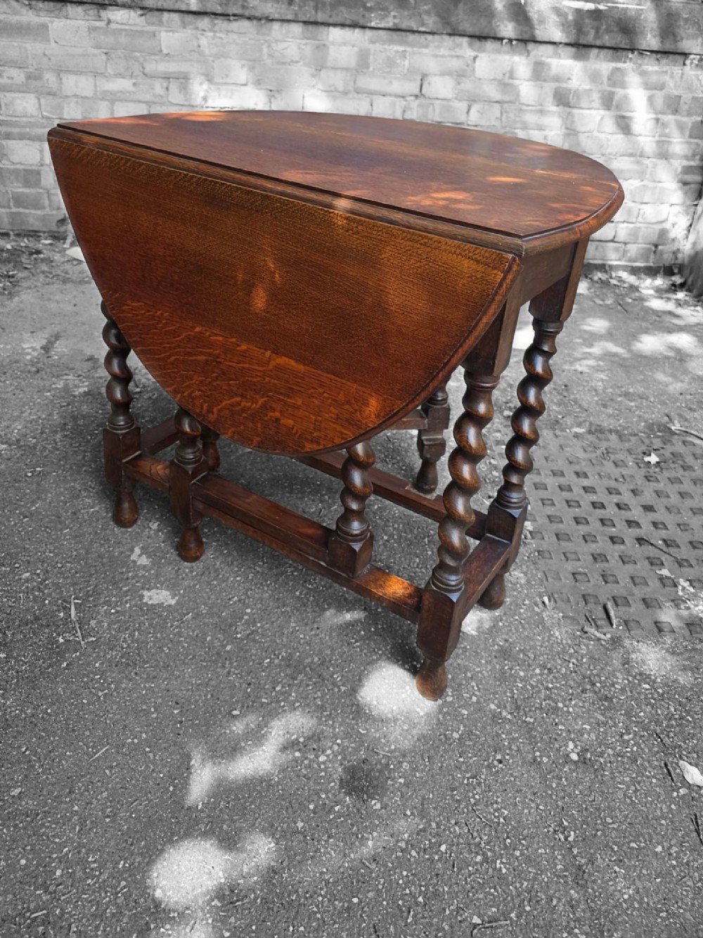 A medium brown oak drop-leaf gateleg table with barley twist legs standing folded on a paved surface.