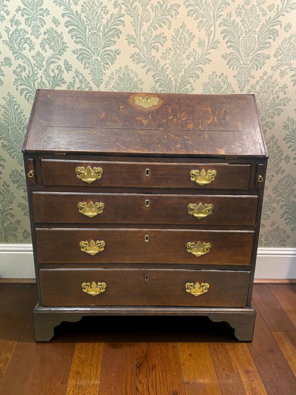 An antique dark oak fall-front writing bureau featuring four horizontal drawers with decorative brass handles and a sloped drop-down lid.