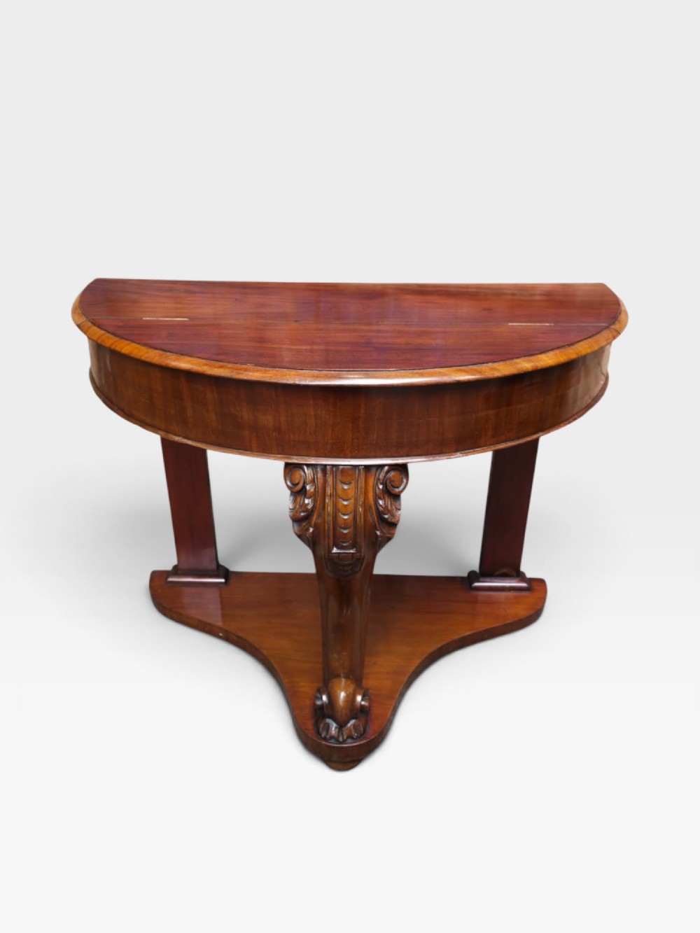 A William IV mahogany demi-lune console table featuring a semicircular fold-over top, a central carved scroll leg, and a shaped plinth base.