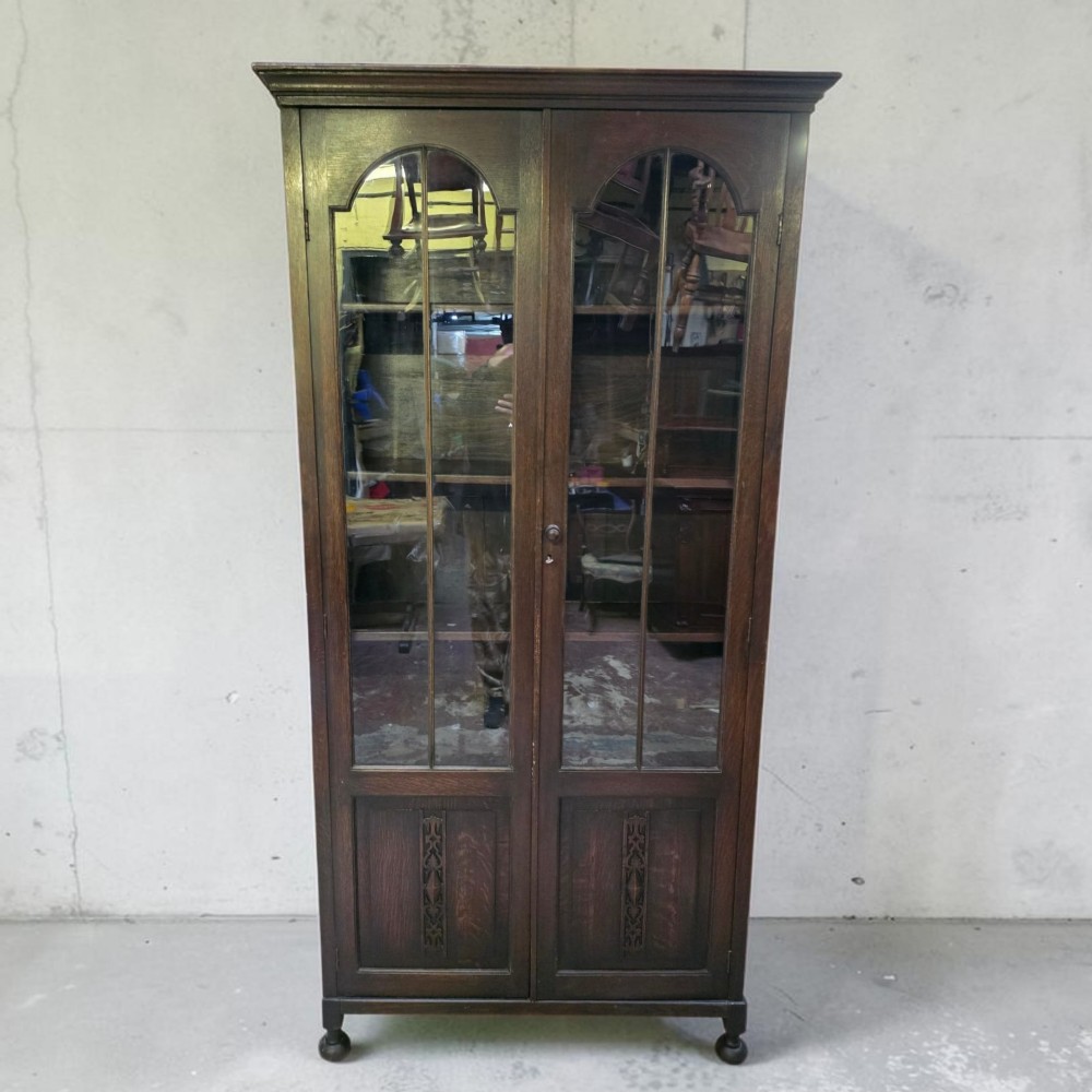 An antique dark oak Arts and Crafts glazed bookcase featuring two tall glazed doors with arched panels above carved lower sections, supported on turned bun feet.