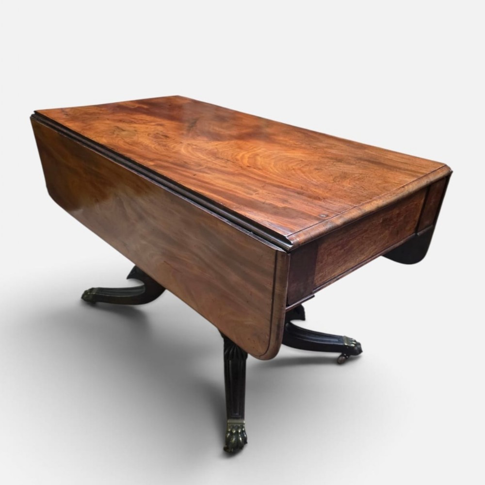 A Regency mahogany drop leaf dining table featuring a flame mahogany top, two hinged side flaps, a central pedestal base, and reeded sabre legs ending in brass claw feet with castors.