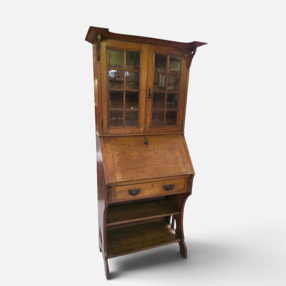 An Arts and Crafts oak bureau bookcase featuring a twin-door glazed upper cabinet, a slant-front writing slope, a single drawer, and open shelving below.