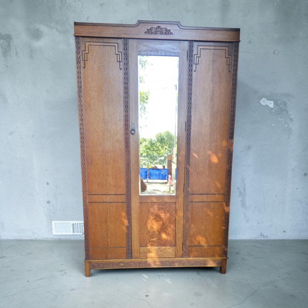 An antique 1920s Art Deco oak wardrobe featuring a central mirrored door, geometric inlay detailing, and carved accents.