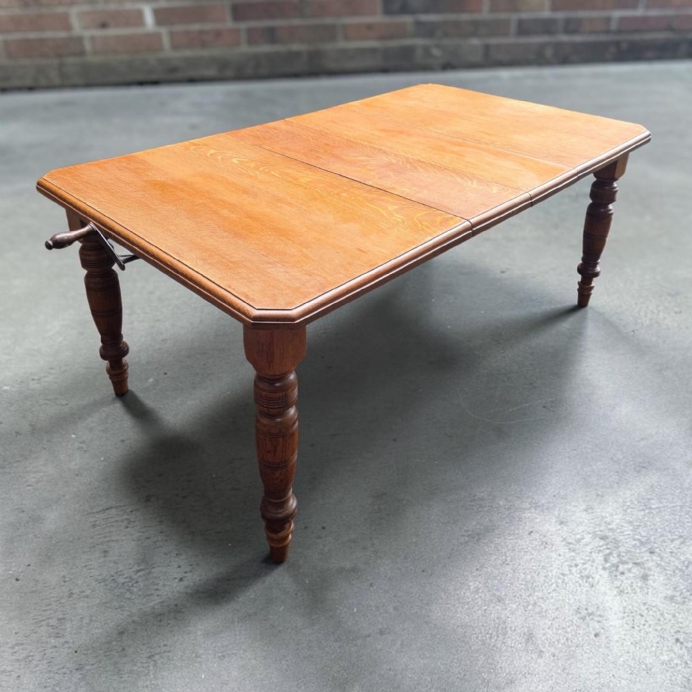 A late Victorian oak extending dining table featuring turned legs, clipped rectangular corners, and an integrated metal winding handle mechanism.