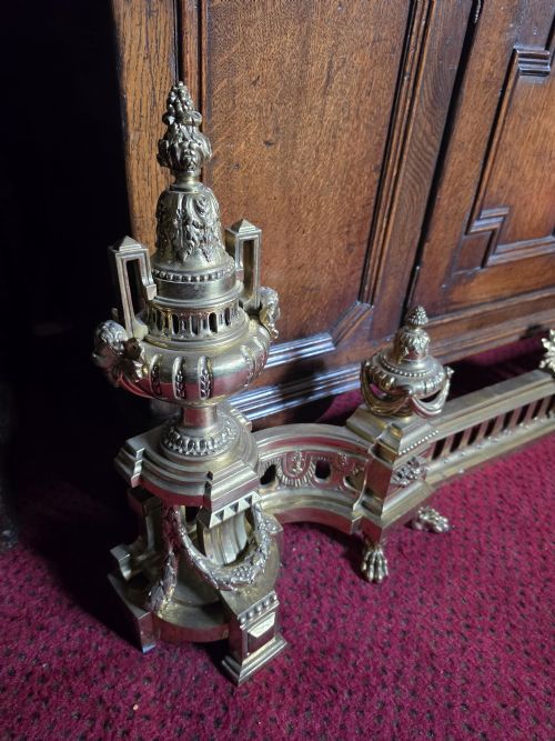 fig 1. An ornate Louis XVI style brass fireplace fender featuring urn finials, classical mask details, and a pierced decorative rail.
