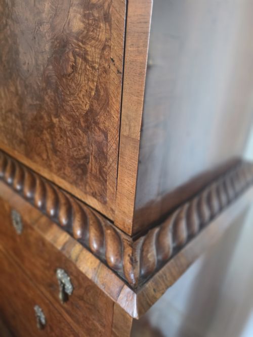 fig 1. An early eighteenth-century English burr walnut escritoire featuring a carved architectural crest, fall-front writing surface, brass drop handles, and a chest of drawers base resting on bracket feet.