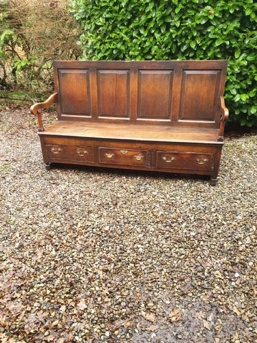 fig 1. An 18th-century dark oak box settle with a four-panelled back, curved armrests, and three base drawers with brass handles.