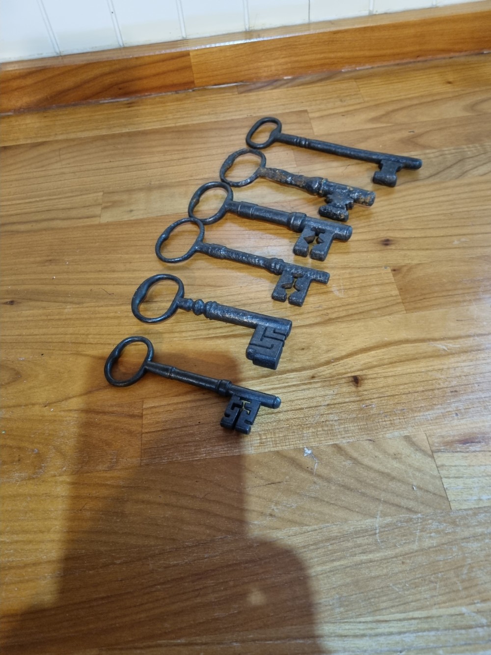 A set of six antique dark cast iron keys with rounded bows and notched bits lying in a row. View VI