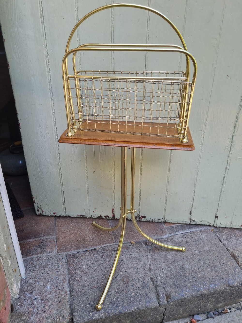 A tall standing brass magazine rack featuring a wire mesh basket, a polished mahogany base, and a three-legged brass tripod stand. View VII