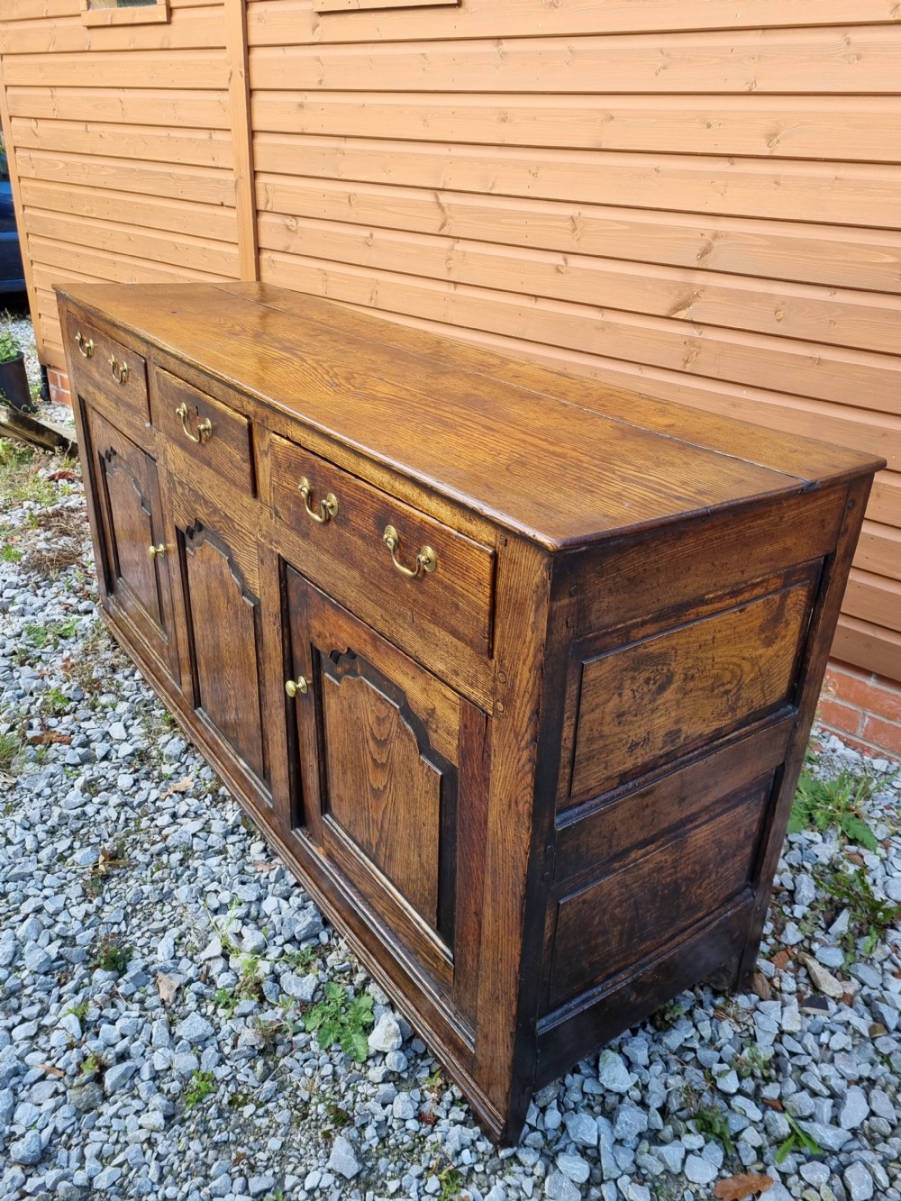 A mid-18th century dark oak dresser base featuring three top drawers over three fielded panel cupboard doors with brass swan neck handles. View II