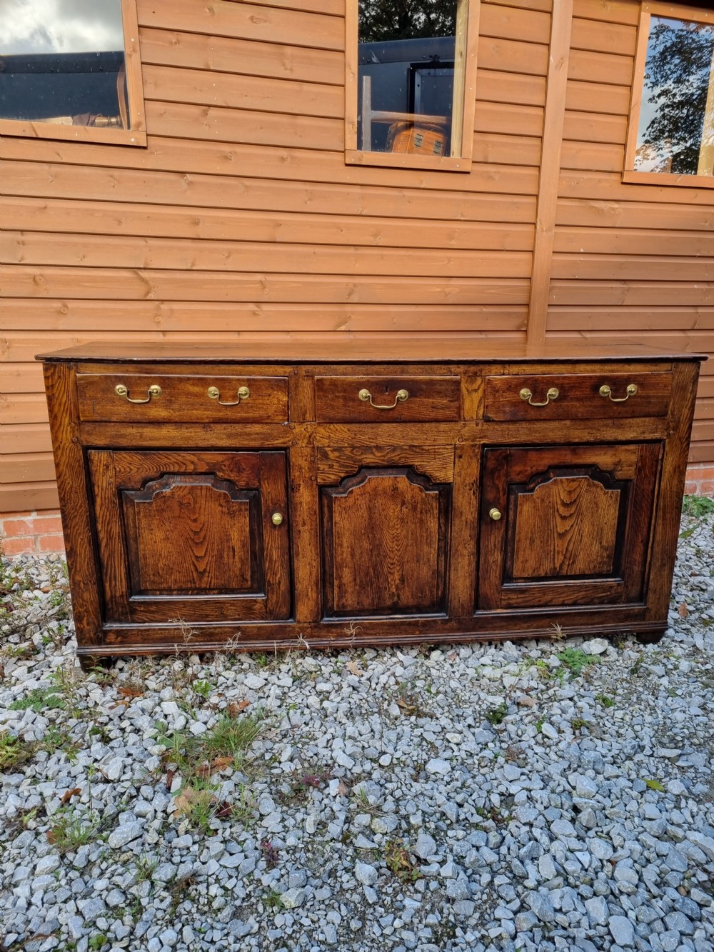 A mid-18th century dark oak dresser base featuring three top drawers over three fielded panel cupboard doors with brass swan neck handles. View VI