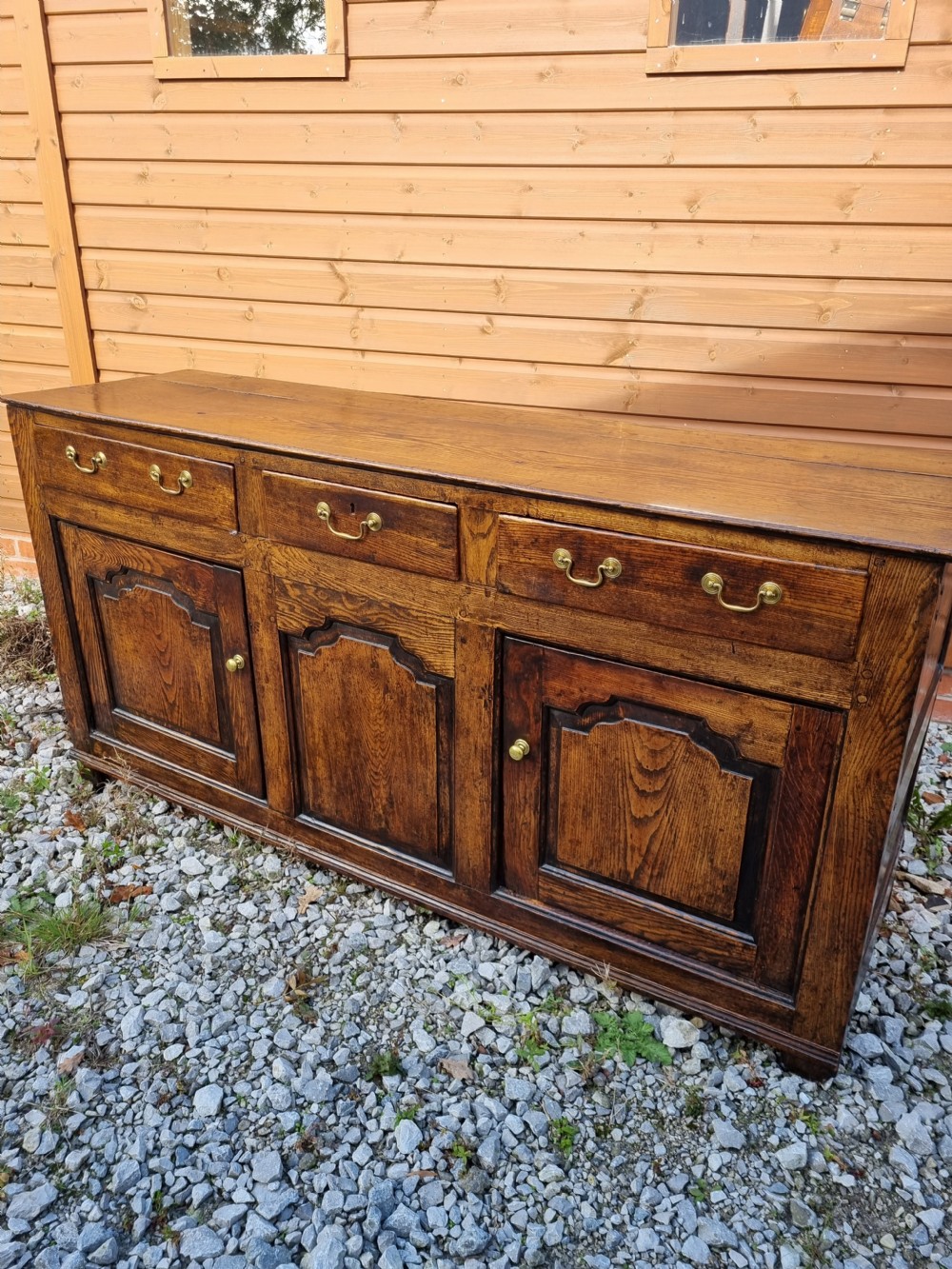 A mid-18th century dark oak dresser base featuring three top drawers over three fielded panel cupboard doors with brass swan neck handles. View VIII